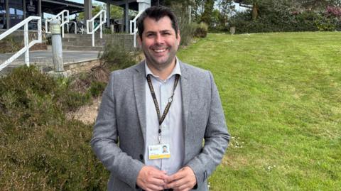 Leigh Frost is smiling as he stands in a garden with County Hall behind him. He has dark hair and brown eyes and is wearing a grey jacket and a grey and white striped shirt and a lanyard with his Cornwall Council identification on it. 