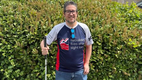 Wayne stands in front of a green hedge, smiling at the camera. He is holding a cane and is wearing a blue top that says 'rebuilding lives after sight loss'.