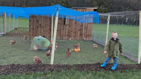 A boy standing in front of a chicken coop. He is wearing a green coat, jeans and blue wellington boots.
