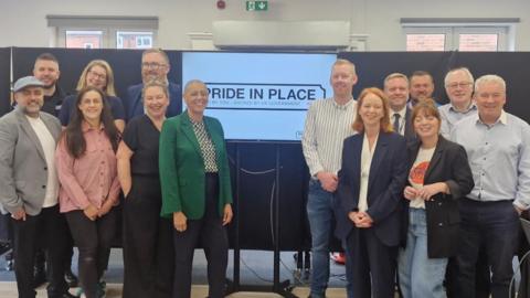 The Pride in Place board: seven women and eight men standing in front of boards and a white screen that reads: Pride in Place