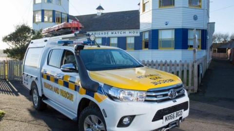 A white and yellow search and rescue vehicle parked outside Tynemouth Volunteer Life Brigade watch house. The building is made of wood and painted white, blue and yellow.