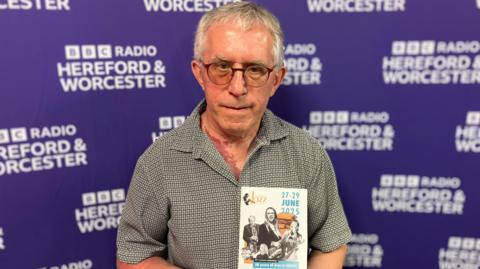 A man stands in front of purple board holding a leaflet.