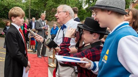 People of all ages - young and old - are dressed up for the film festival. One child is being interviewed on the red carpet by an older man and is wearing a suit and tie, while another person wearing a hat is taking a photo. A third young person stands looking with a notepad and pen in his hand. 