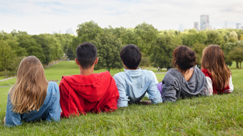 Five boys and girls lie on a hill looking down over parkland. Their backs are to the camera. They have hooded tops and/or casual clothing on. Trees line part of the park with tower blocks visible in the background on the right-hand side.