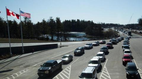 Cars wait in three lanes at the US-Canada border with the flags of the two countries seen on the left
