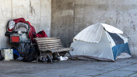 A tent and a supermarket trolley loaded with posessions are either side of a bench on an urban street.