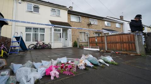 A cordon outside the front of the property with a police car parked in the road. Flower tributes have been left on the driveway of the house. 