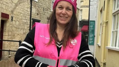 Chrissie Lowery. She has long dark hair and is wearing a bright pink beanie and a black and white stripe jumper under a pink high-vis vest. It has the Safe Space logo on one side and the words Night Angels on the other. Chrissie is looking directly at the camera and smiling. 