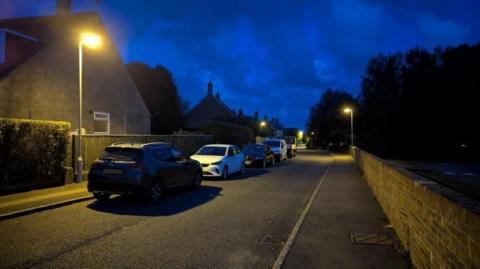 A street at night. There are houses on the left hand side of the street with cars parked alongside them. On the right hand side of the road is a brick wall and then some trees. There are yellow street lights at regular intervals.