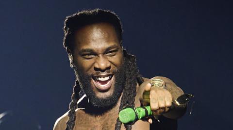 Burna Boy pointing with his finger, holding a green microphone