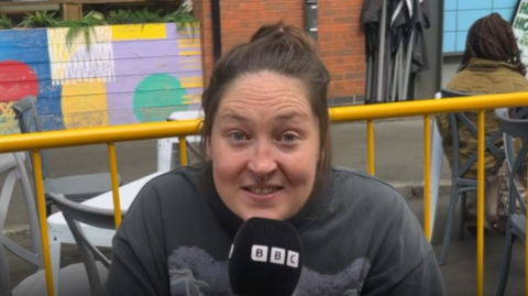 A Manchester woman sitting outside at a restaurant wears a grey t-shirt and talks into a 鶹Լ microphone