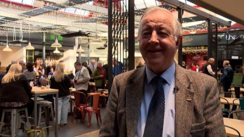 Ailig Greumach na sheasamh sa mhargaidh an Inbhir Nis.