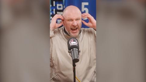 Steve Bracknall holds his hands to his ear with an angry expression on his face