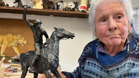 A brass sculpture of a polo player on a running horse sits next to Jan, who is in her 80s, in her studio at home in Stawell. 