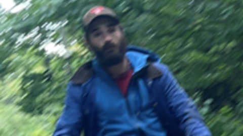 A blurry image of a man in a blue anorak with a red t-shirt underneath. He is white with a black beard, heavy black eyebrows and a brown baseball cap with a red logo on it.  