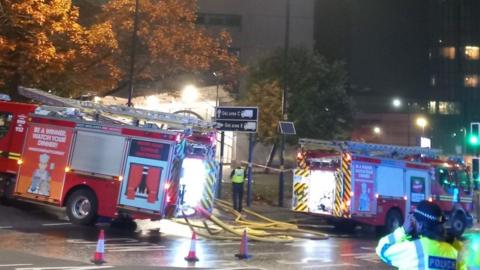 Two fire engines parked outside a tower block with hoses coming out of them. Trees are in front of the building on the left and a police officer is in the foreground. 