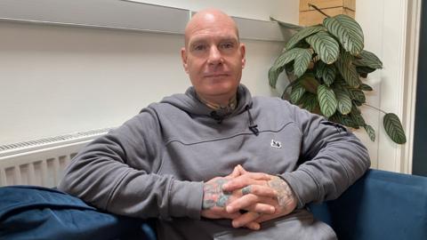 A man in a grey hoodie sits on a sofa. He interlocks the fingers of each hand, holding them above his stomach. He has a shaved head and tattoos on his neck and hands. There is a plant and a radiator behind him. 