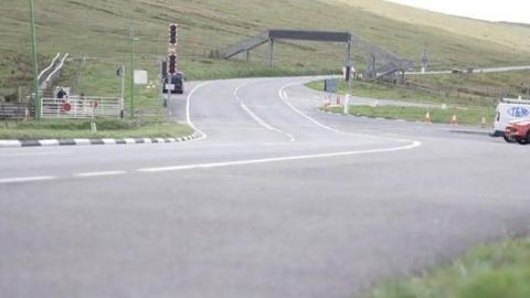 Mountain Road at the Bungalow. The carriageway snakes off into the distance under a metal footbridge over the road. There are electric tram signals on the left and a sloping hill in the distance.