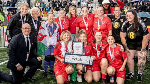 Group of women standing in a football pitch, they are holding a trophy and there is seating in the background there are also flags in the back