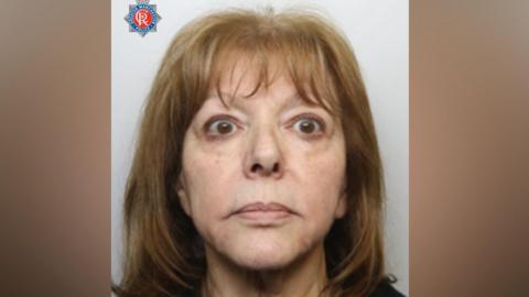A woman with shoulder-length dark blonde hair is looking at the camera in a police mugshot. There is a Greater Manchester Police logo in the top left hand corner.