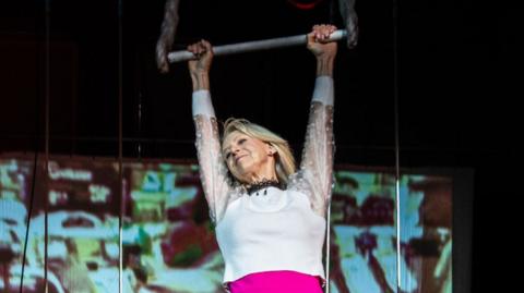 A woman hangs from a trapeze with both hands  - wearing a sparkly white top and pink leotard. She is smiling and looking to one side.