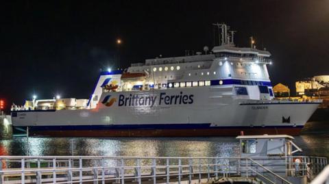 A white Brittany Ferries boat called Islander. It is night time and the vessel is lit up with lights around its decks. The boat is docked in a port.