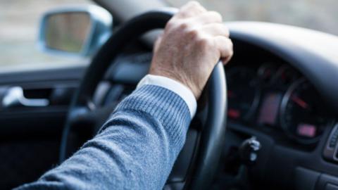 An elderly man's hand on a steering wheel in a car.