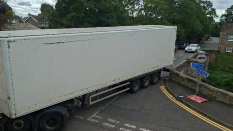 A large lorry with a white trailer squeezes across a narrow stone bridge, with trees and houses in the background. A sign advertises a 7.5-tonne weight limit
