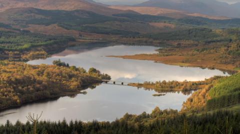 A view across Loch Garry on a fine autumn day. The surface of the loch is flat calm and trees along its shore are in autumn colour. The loch resembles the shape of mainland Scotland.