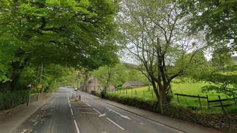 A semi-rural road with no cars on it. It is flanked by footpaths, a dry stone wall, trees and grass. There is a pedestrian crossing island in the middle distance.