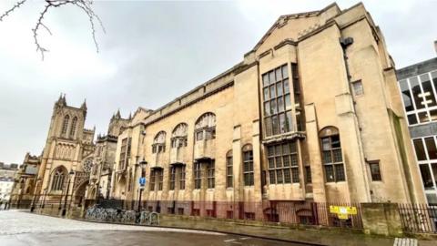 The Bristol Central Library building fills the frame. It is a beige building with old window frames. Bikes are parked outside.