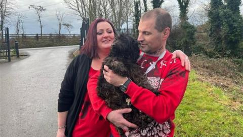 A woman with dark red hair wearing a red top and black cardigan and a man in a red patterned top stand in a park. They are cuddling a dark brown shaggy dog in their arms. 