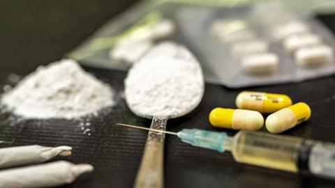 A table with drugs on including a spoon with white powder, a needle, pills and joints.