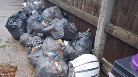 About a dozen black bin bags, containing waste, left on concrete next to a brown, weathered wooden fence. An old white lampshade can also be seen.