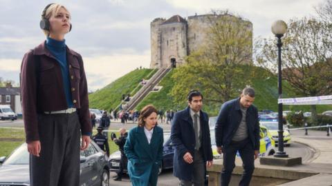 A blonde woman, wearing headphones, looks up towards a building, as three detectives climb steps to the side of her. In the background is Clifford's Tower in York.