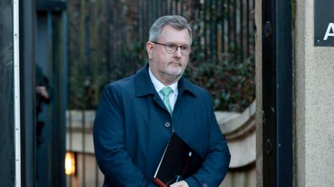 Sir Jeffrey Donaldson, pictured with short grey hair wearing glasses, a white shirt, blue tie and navy jacket. He is holding a notepad. 