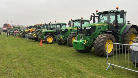 A long line of green tractors parked on grass with some orange bollards nearly and people at the end of the field. The sky is grey.