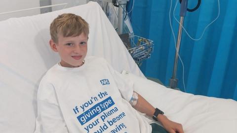 George, who has blonde hair, smiles at the camera from a hospital bed. He is wearing a large white t-shirt with a message about giving plasma on it in blue lettering. 