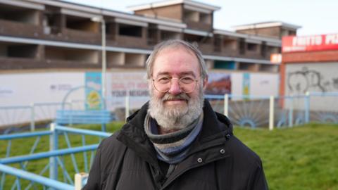 Stephen Spencer smiles at the camera in front of a block of maisonettes and some grass. He wears a black coat and a striped scarf. He has a grey beard and hair.