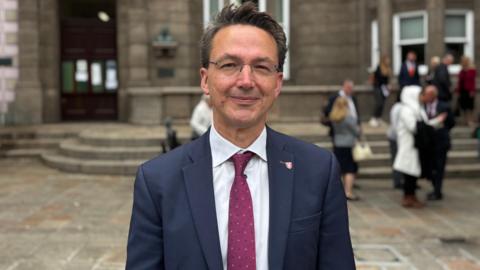Kirsten Morel is standing in front of the stone clad Royal Courts building. He is dressed in a navy jacket with a burgundy coloured tie. He has short dark hair and is wearing glasses.