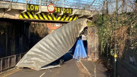 A damaged railway bridge with a piece of metal hanging down onto the road below and the words "low bridge" in large yellow lettering.