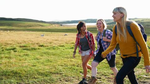 Three female hikers laugh as they walk in an idyllic summer countryside.
