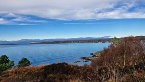 Blue sky with white clouds over water with brown shrubs and scrub land in the foreground 
