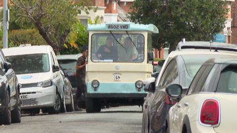 Small vintage blue and cream milk delivery truck labeled "Parker Dairies" driving down a residential street with parked cars and a person nearby.