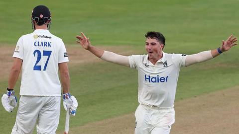 Warwickshire fast bowler Michael Booth celebrates taking the wicket of Sussex's Tom Clark at Edgbaston. He is running towards his team-mates who are out of the picture with his arms out stretched. The batter has his back to the viewer with his name and the number 27 on his shirt.
