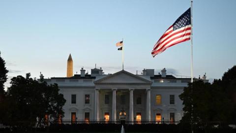 A view of the White House building from outside. An American flag flies in front of it, while the sun is setting.