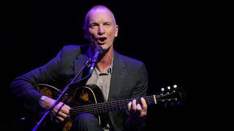 Sting performing at the Baltic Centre for Contemporary Art. He has a shaved head and is wearing a grey suit and shirt. He is sitting down as he sings and plays a guitar next to a microphone.