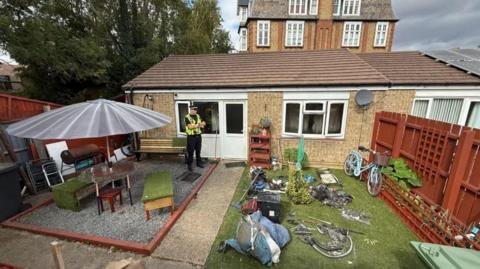 The back garden of the bungalow - with a green lawn and wooden garden furniture. A police officer is in black trousers and a yellow high-vis jacket standing by the backdoor.
