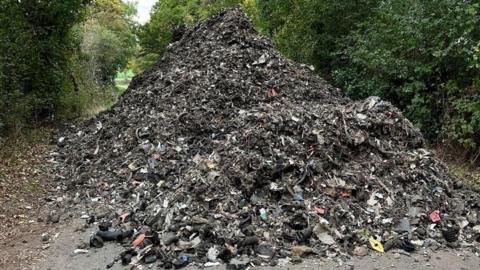 A large pile of rubbish blocking a country road, with trees in the background.