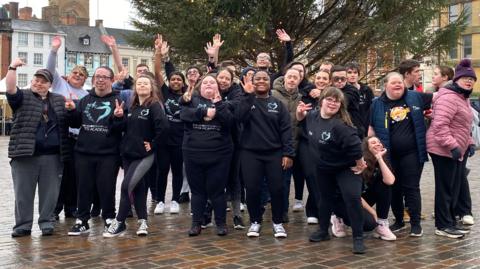 A group of dancers dressed in navy jumpers pose with their hands in the air and smiling at the camera in the middle of a wet town centre. 